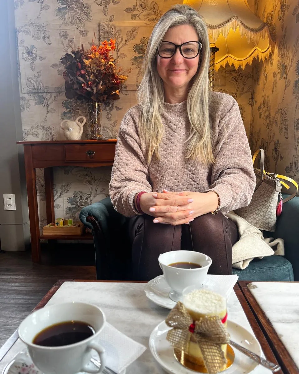 Cíntia sorrindo, sentada numa poltrona em um café, com duas xícaras de café e um doce à frente